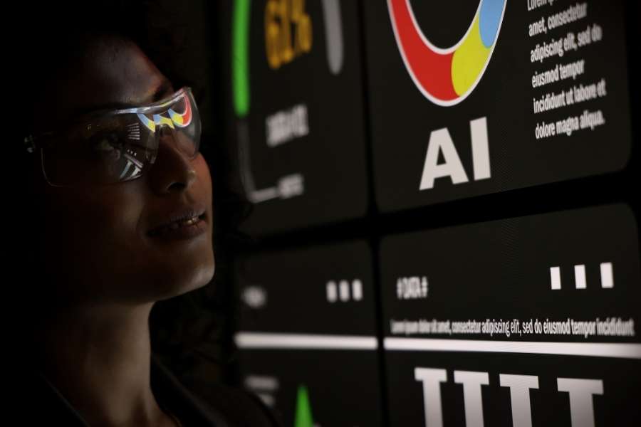 Une analyste portant des lunettes de protection observe un écran de contrôle affichant le mot AI et des graphiques.
